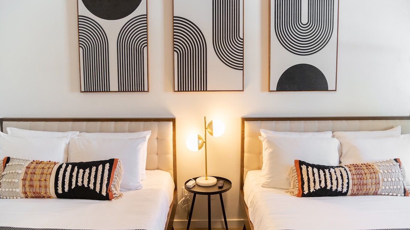 Two beds with white bedding and pillows, black and orange accent pillows, artwork above, and a lamp between them.