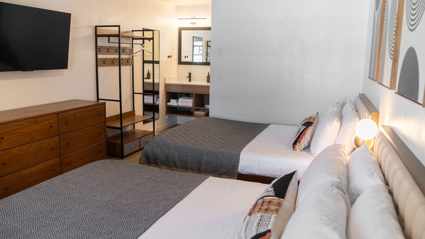 A modern hotel room with two beds, a dresser, and a bathroom visible in the background.