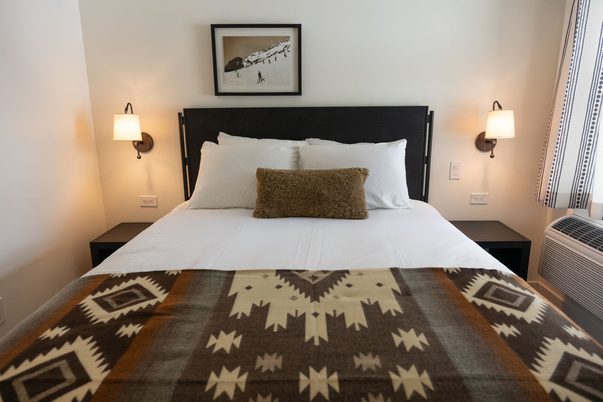 Bedroom with bed, patterned blanket, decorative pillows, wall art, and sconces.