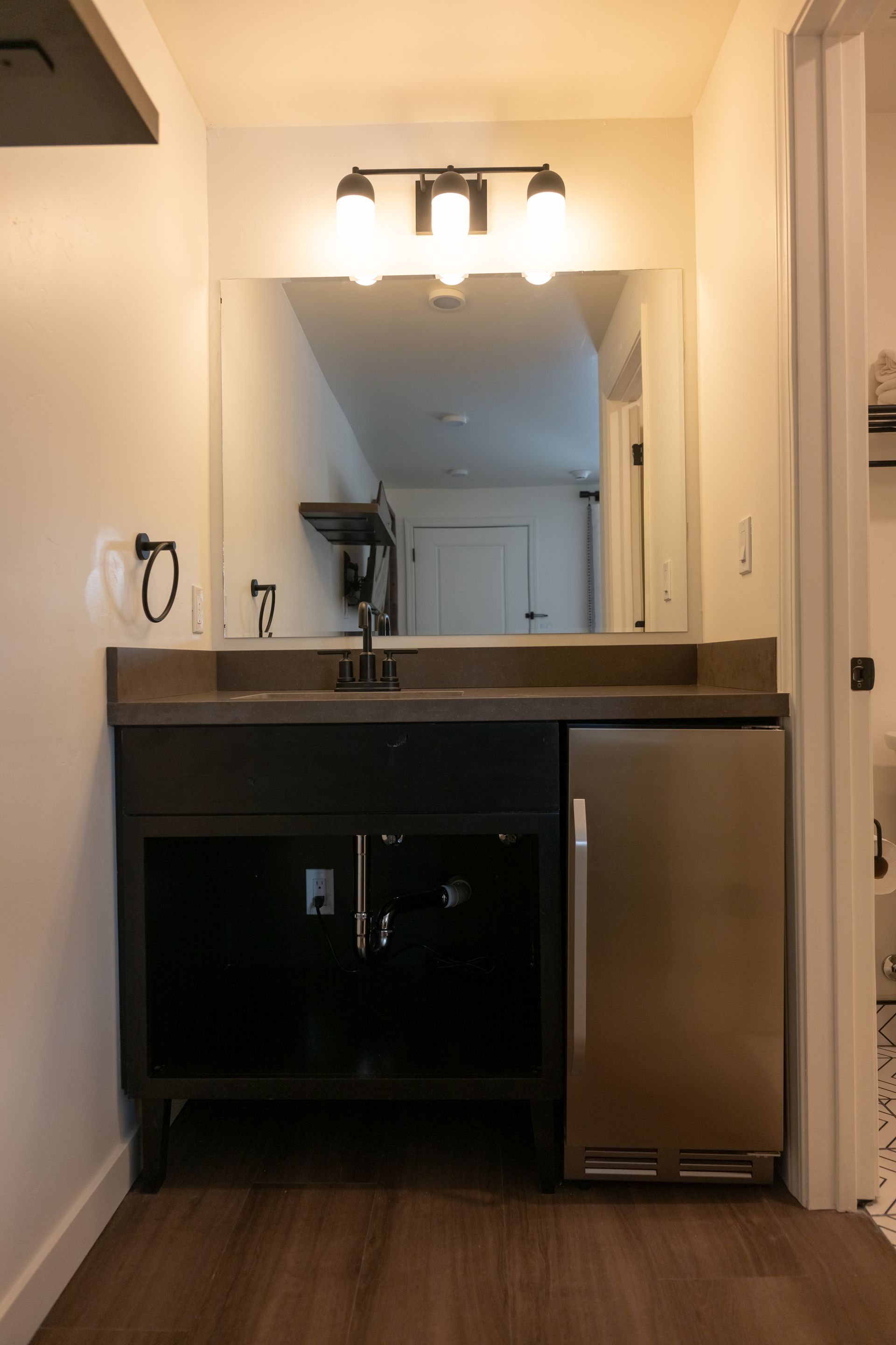 Bathroom vanity with a dark cabinet, mirror, and mini-fridge. The room has white walls and dark wood-look flooring.