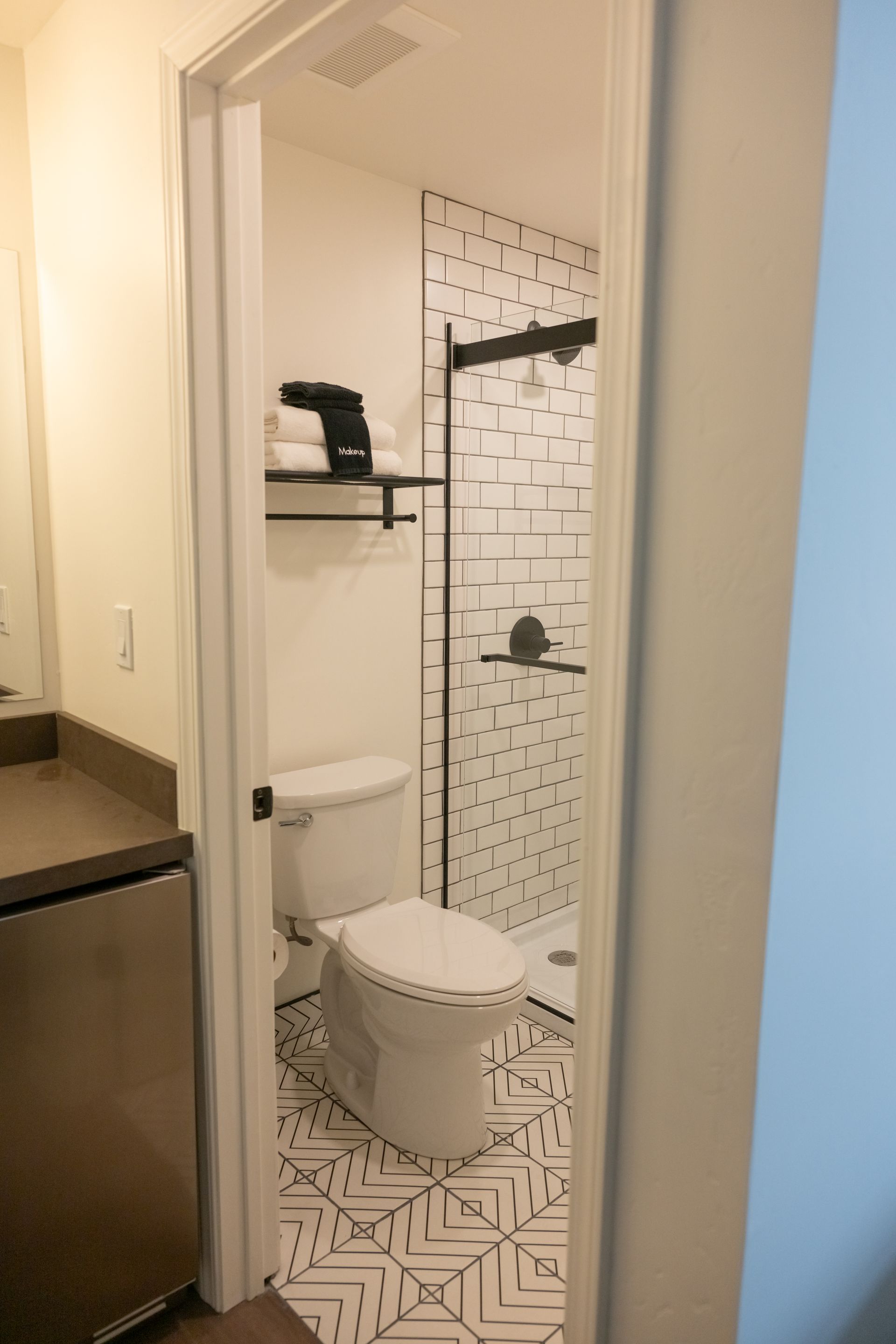 Narrow bathroom with patterned floor, white toilet, black shower fixtures, and towels on a shelf.