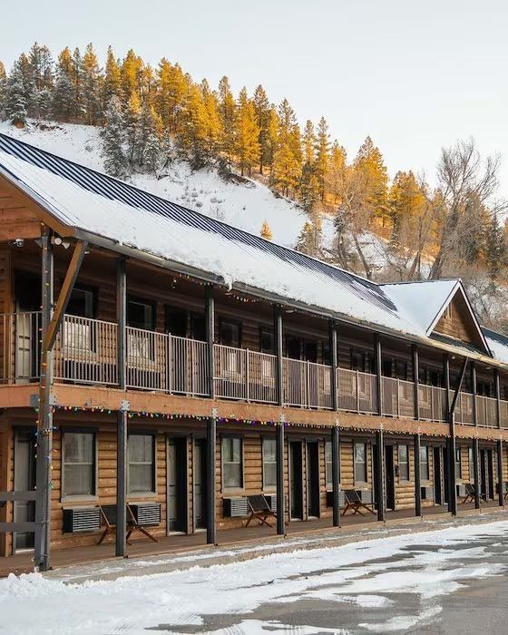 Two-story log cabin motel in winter. Snow on the ground and roof, trees on the hillside.
