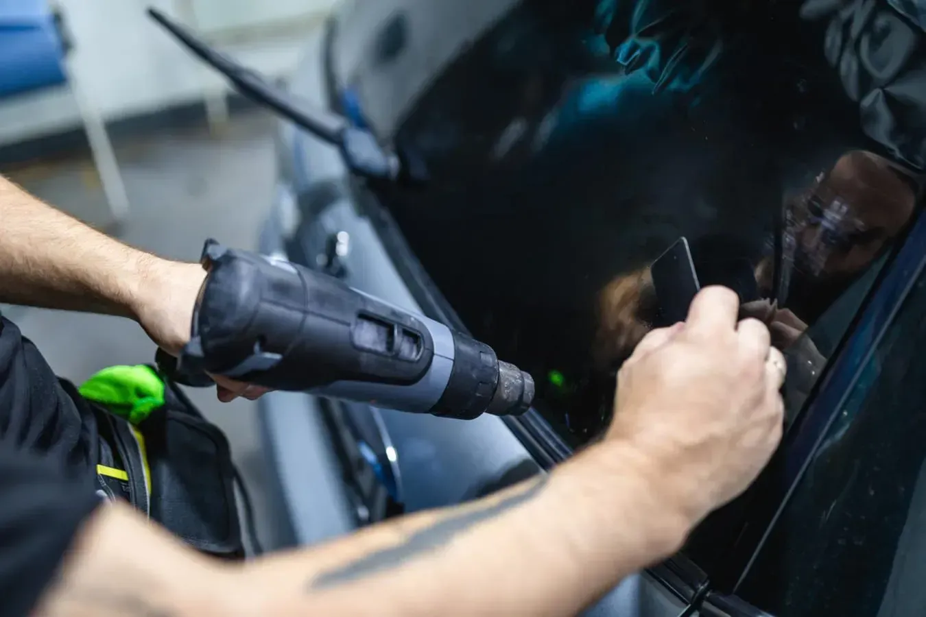 Person applying window tint to a car windshield with a heat gun in a garage.