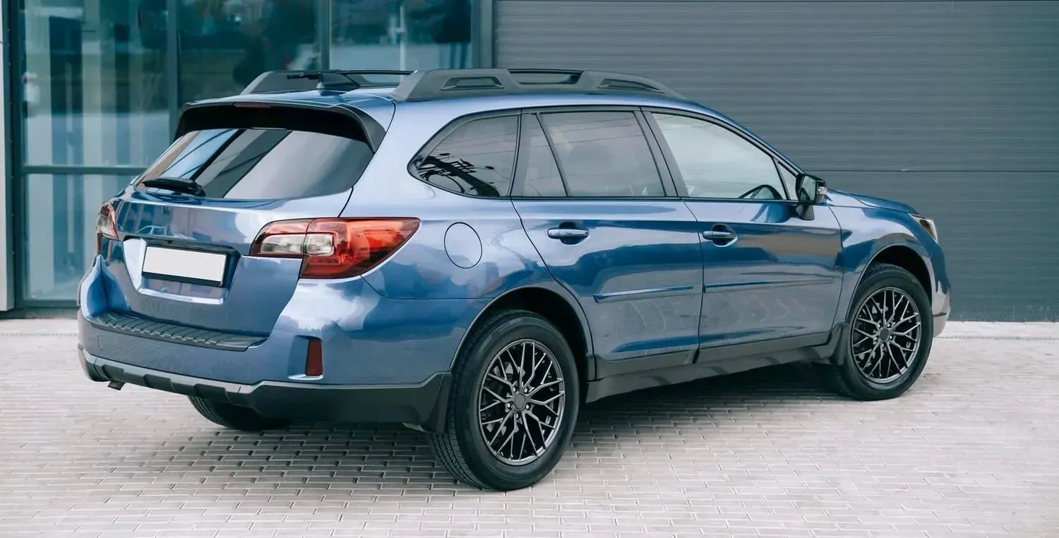 Blue Subaru Outback parked in front of a gray building with black wheels and roof rack.