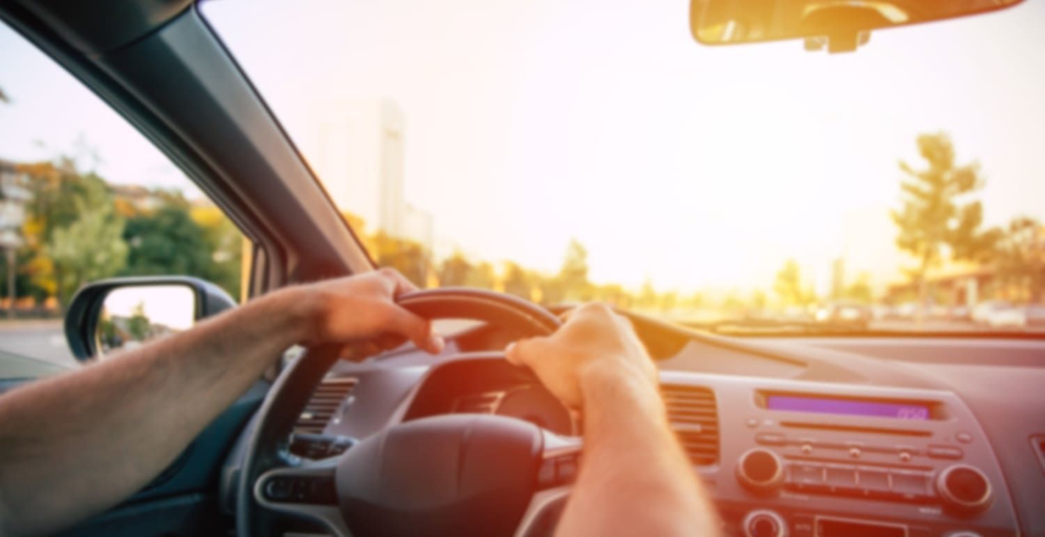 Hands gripping a steering wheel inside a car, driving toward a bright, sunny horizon.