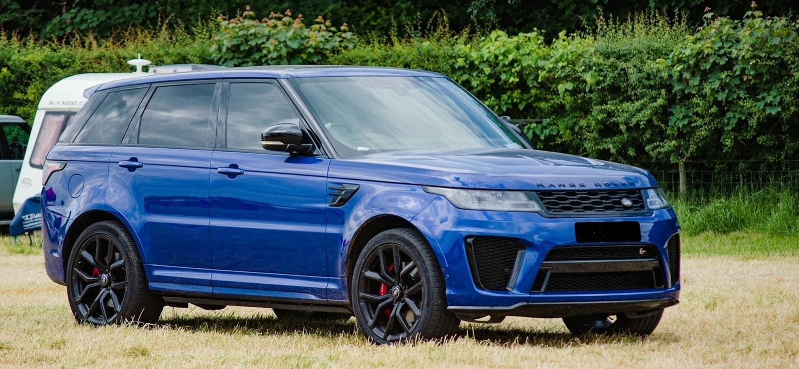 Blue Range Rover SUV parked on grass. Black wheels with red brake calipers. Hedge in background.