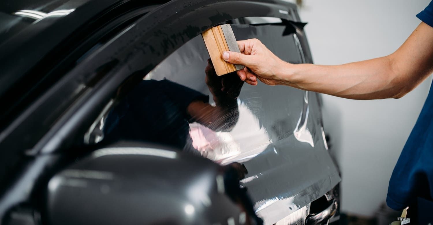 Person applying window tint to a car window with a squeegee in a workshop.