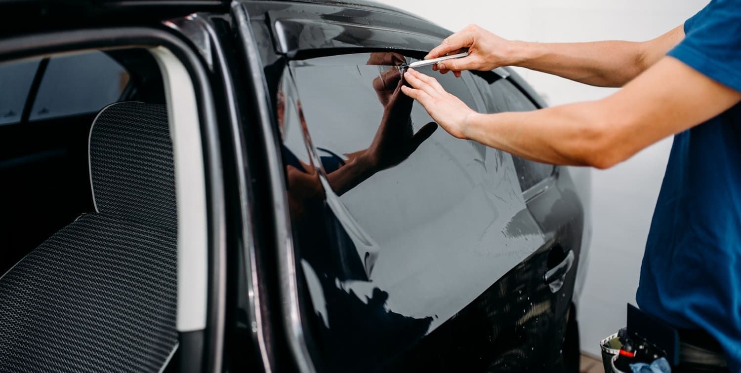 Person applying tint to a black car window with a utility knife in an outdoor setting.