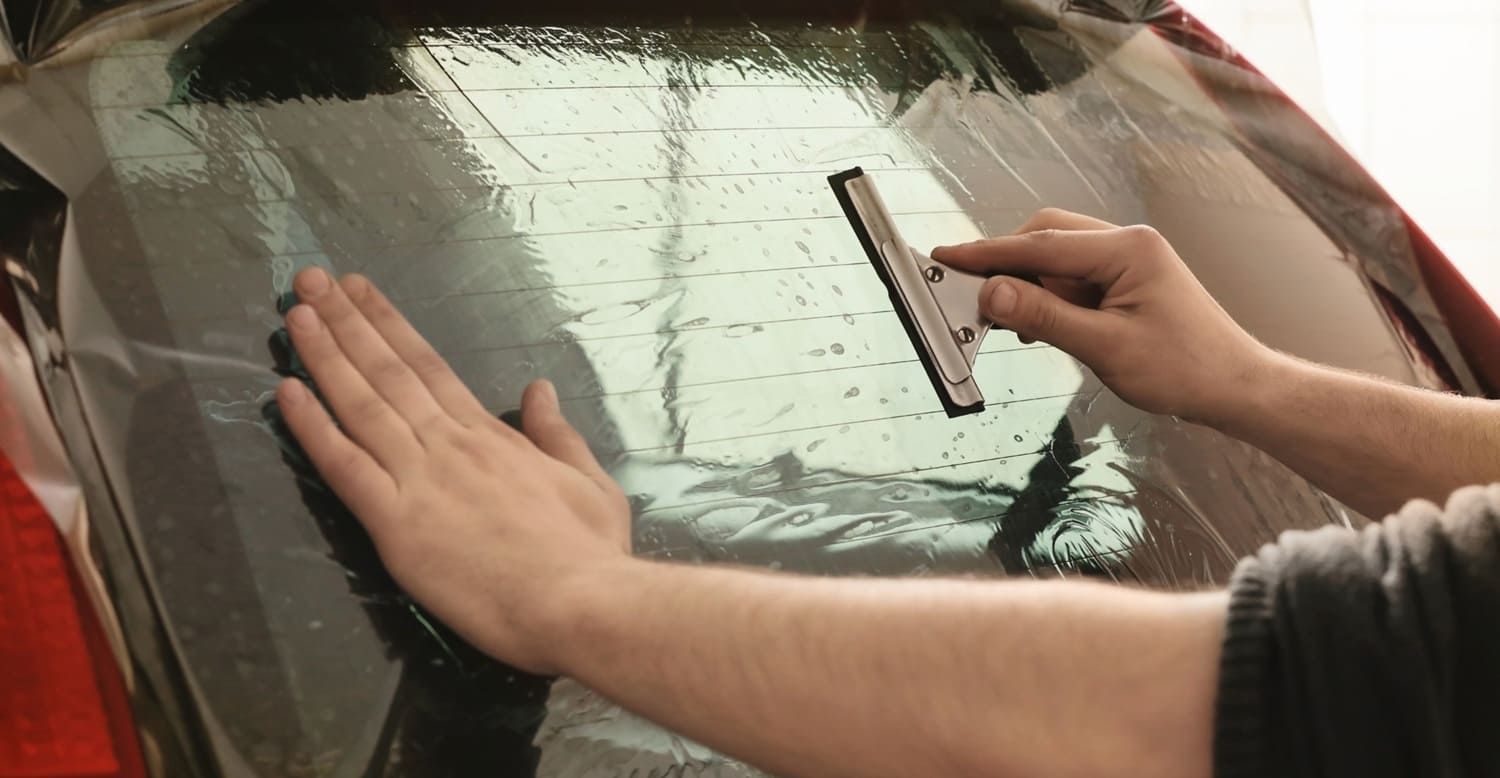 Hands applying window tint to car's rear window with a squeegee.