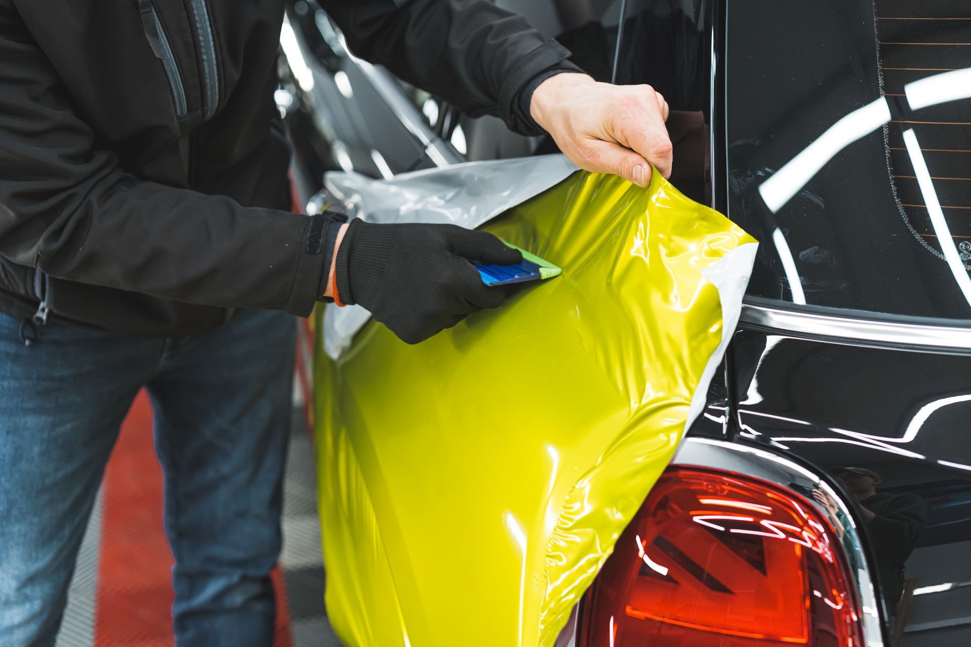 Person wearing gloves applying yellow vinyl wrap to a black car's taillight with a tool.