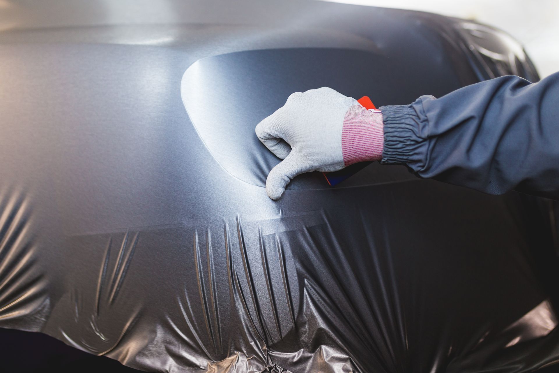 A person in a gray jumpsuit and gloves smooths dark car wrap onto a vehicle hood.