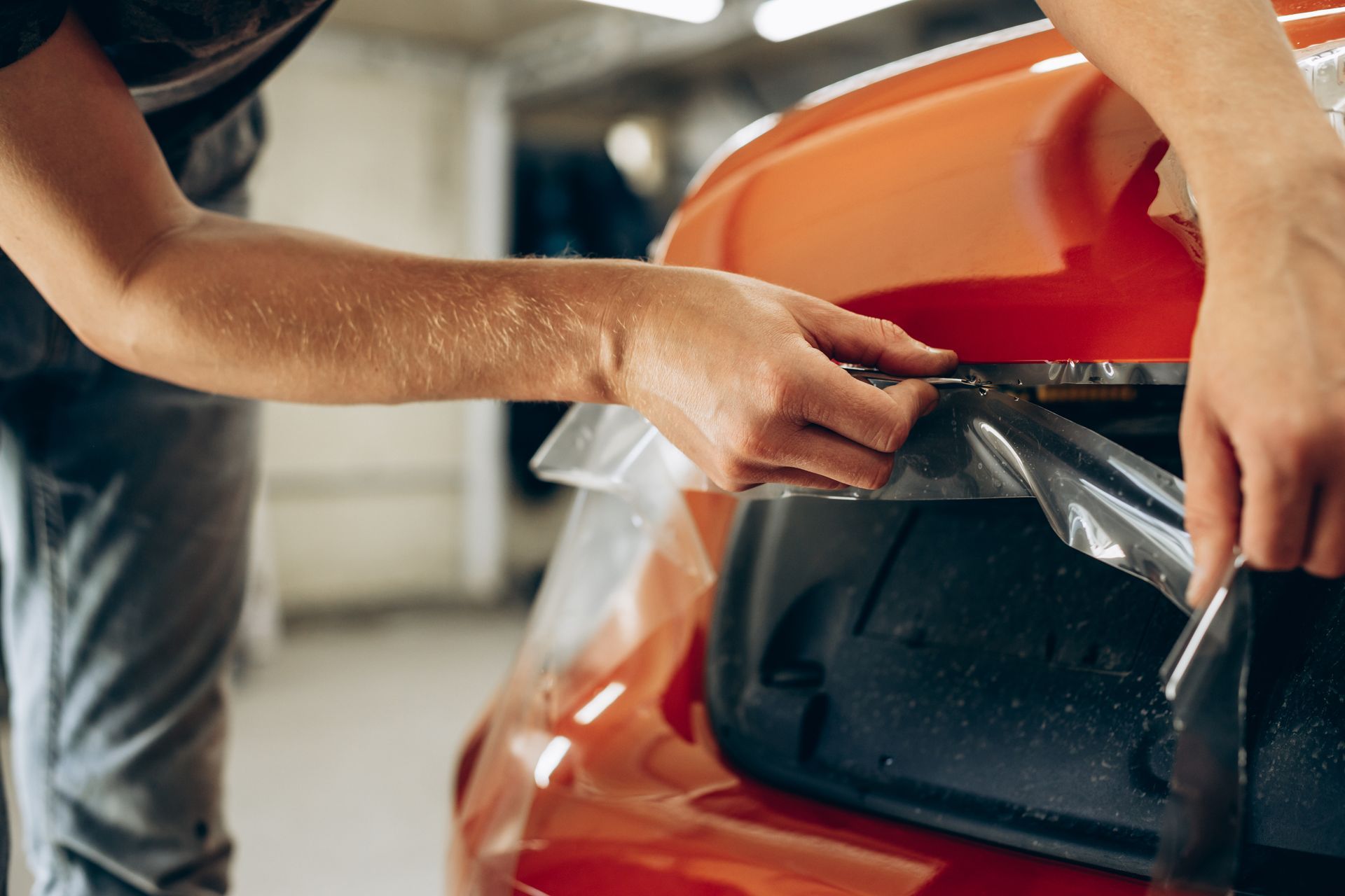 Hands applying clear film to an orange car's taillight in a garage.