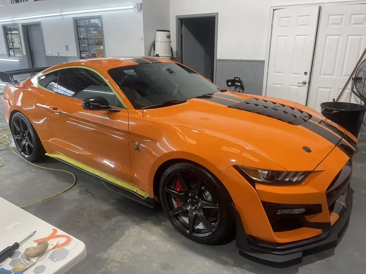 Orange Ford Mustang with black racing stripes in a garage.