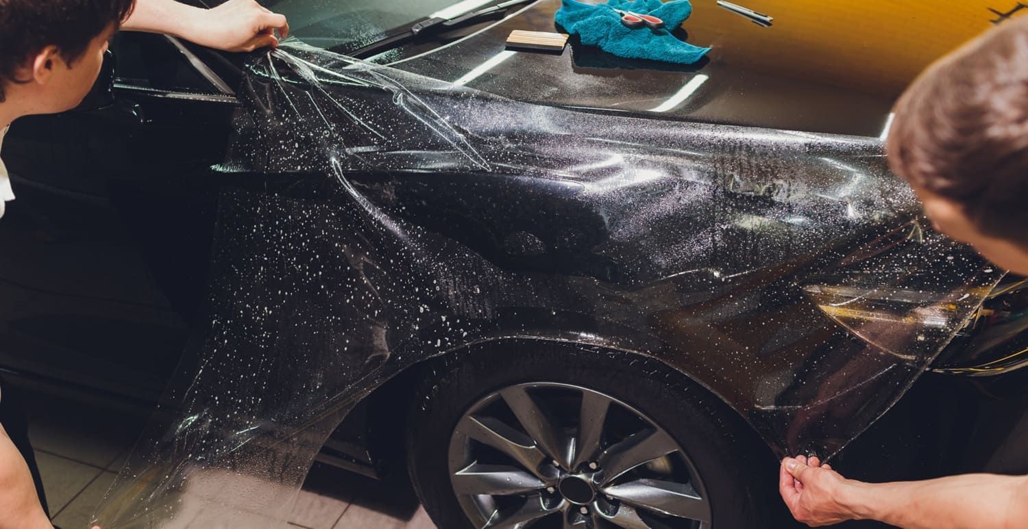 Two people applying a clear protective film to a black car's hood and fender.