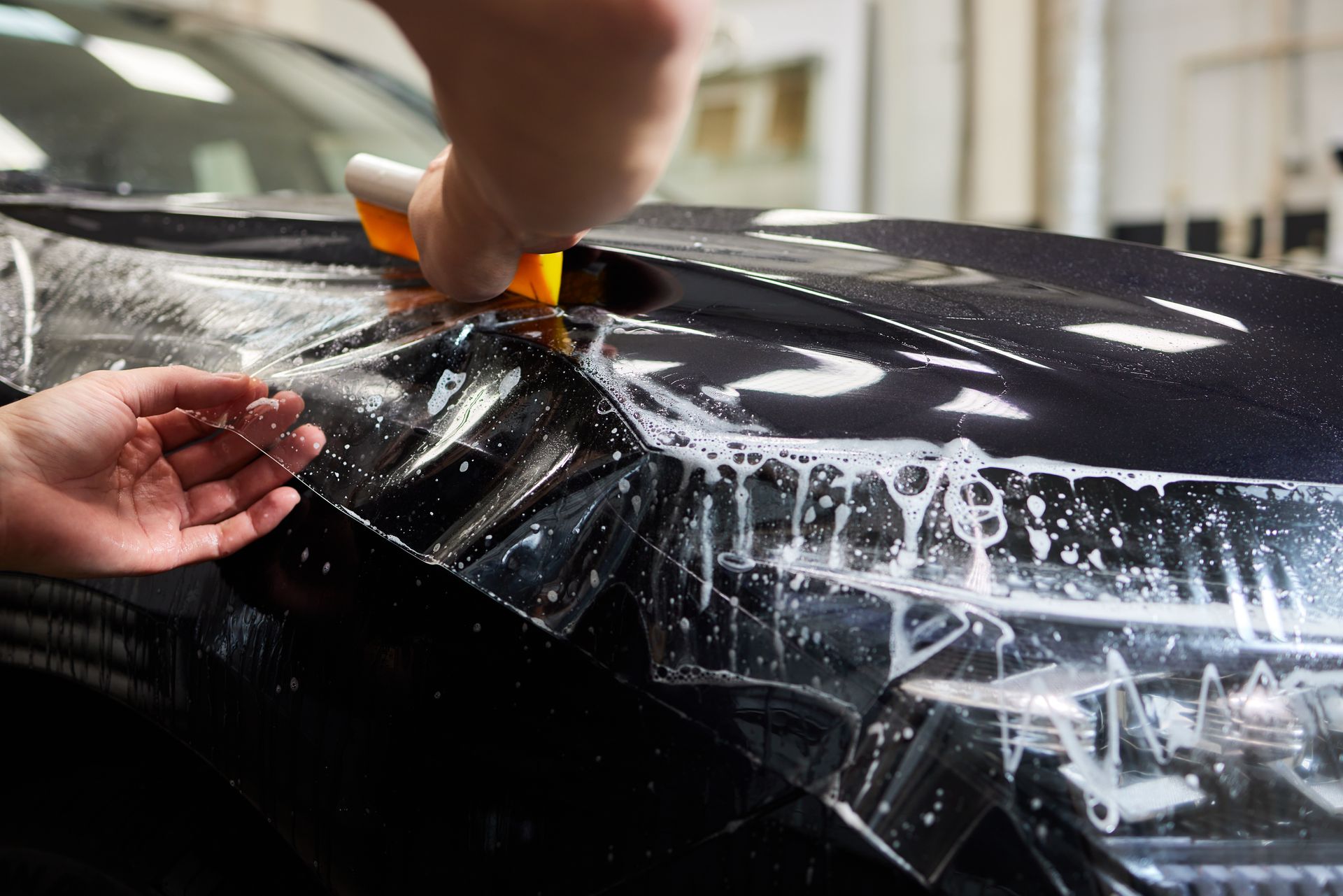 Person applying protective film to a black car's hood, using a squeegee and spray.