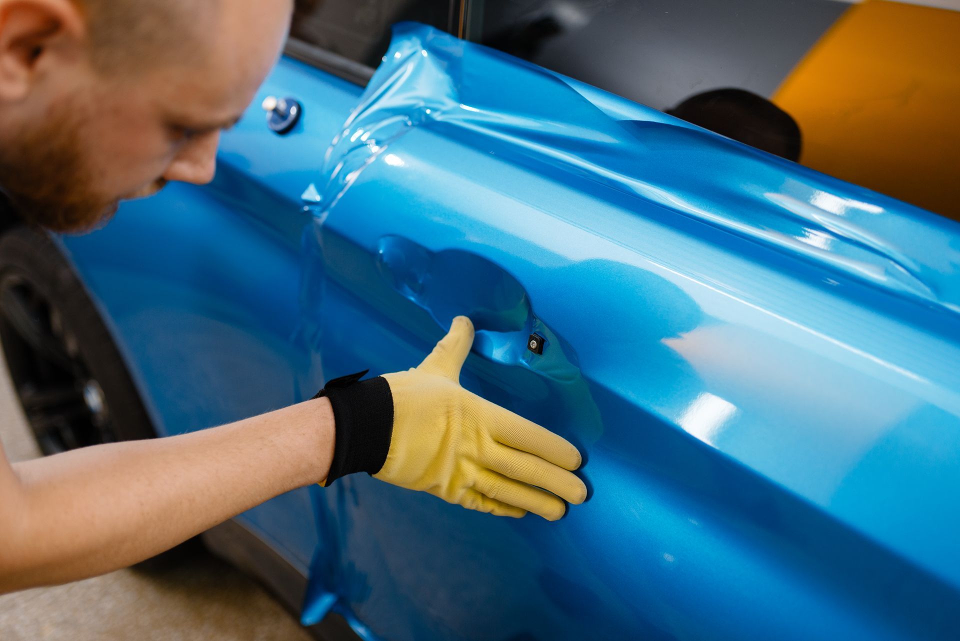 Person wearing yellow gloves applying blue vinyl wrap to a car door.