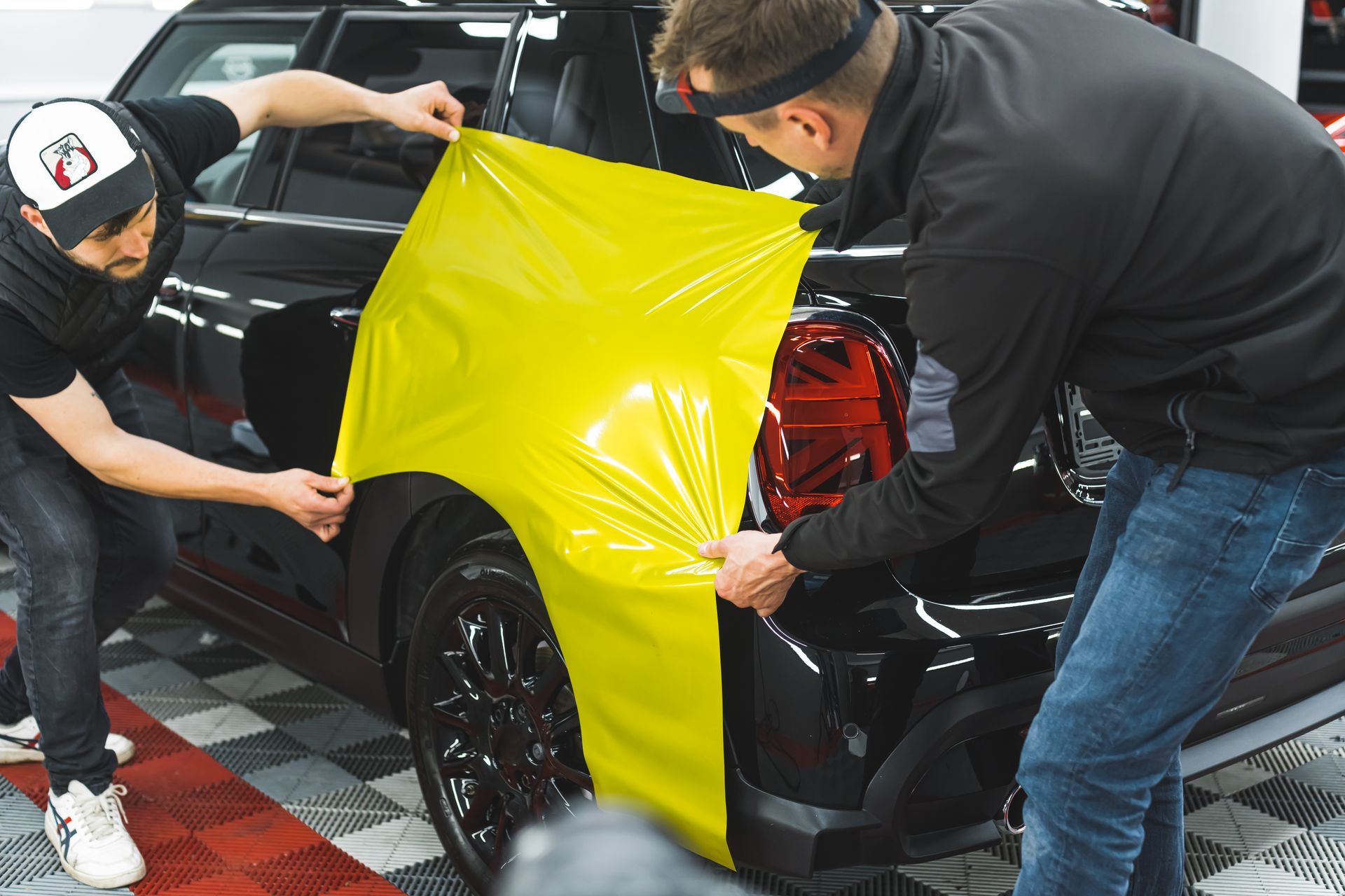 Two people applying a yellow vinyl wrap to the rear of a black car in a garage.
