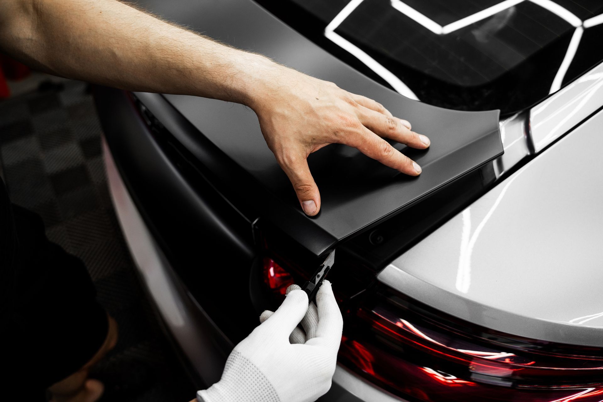 Hands installing a black spoiler on a car's trunk, with one hand wearing a white glove.