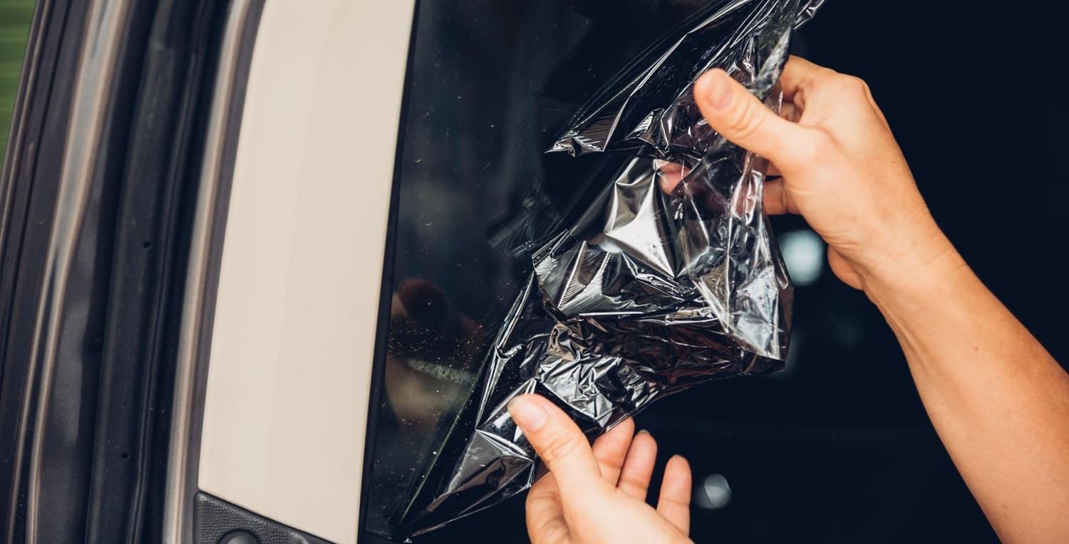 Hands peeling off black window tint from a car window.