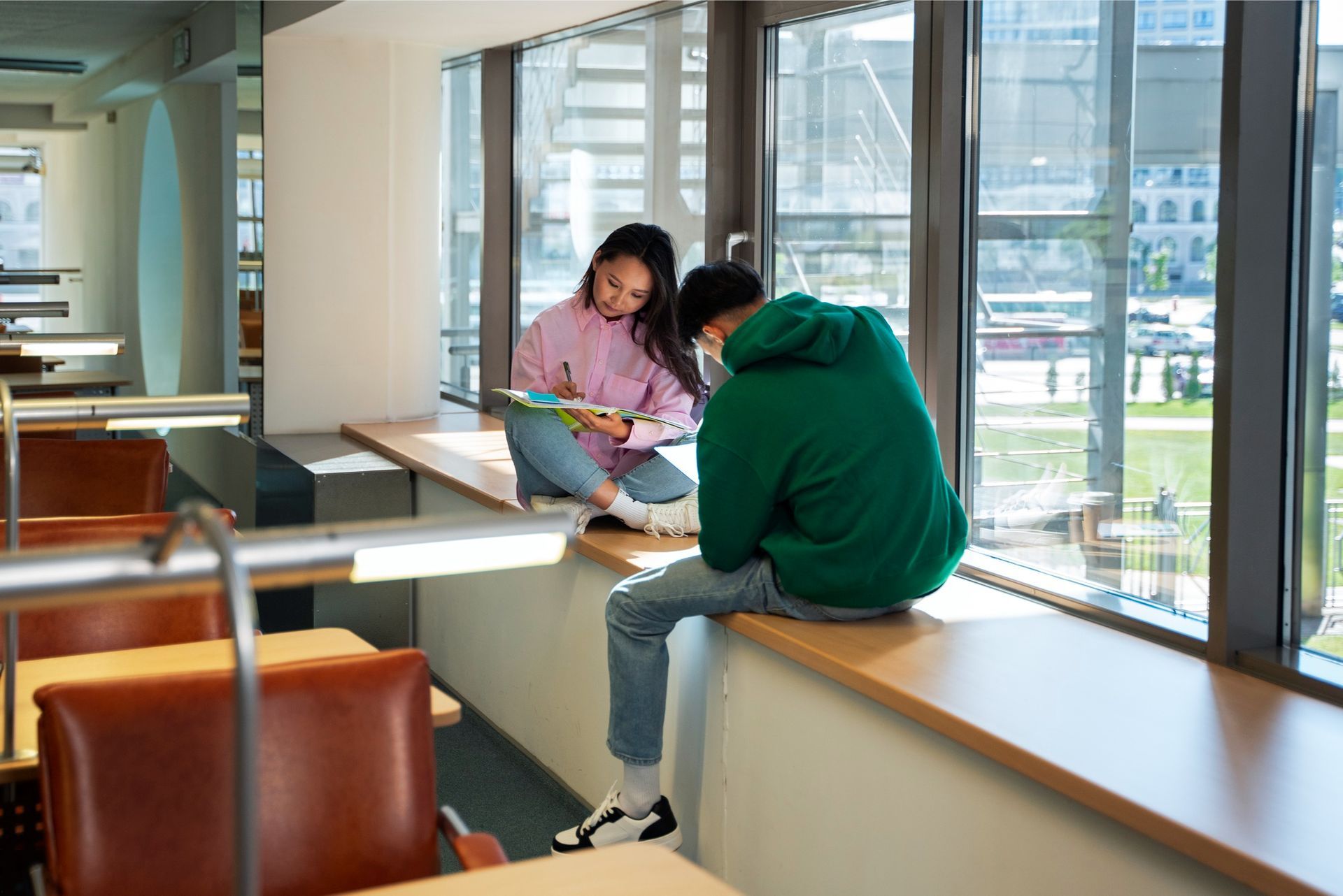 A man and a woman are sitting on a window sill reading books.