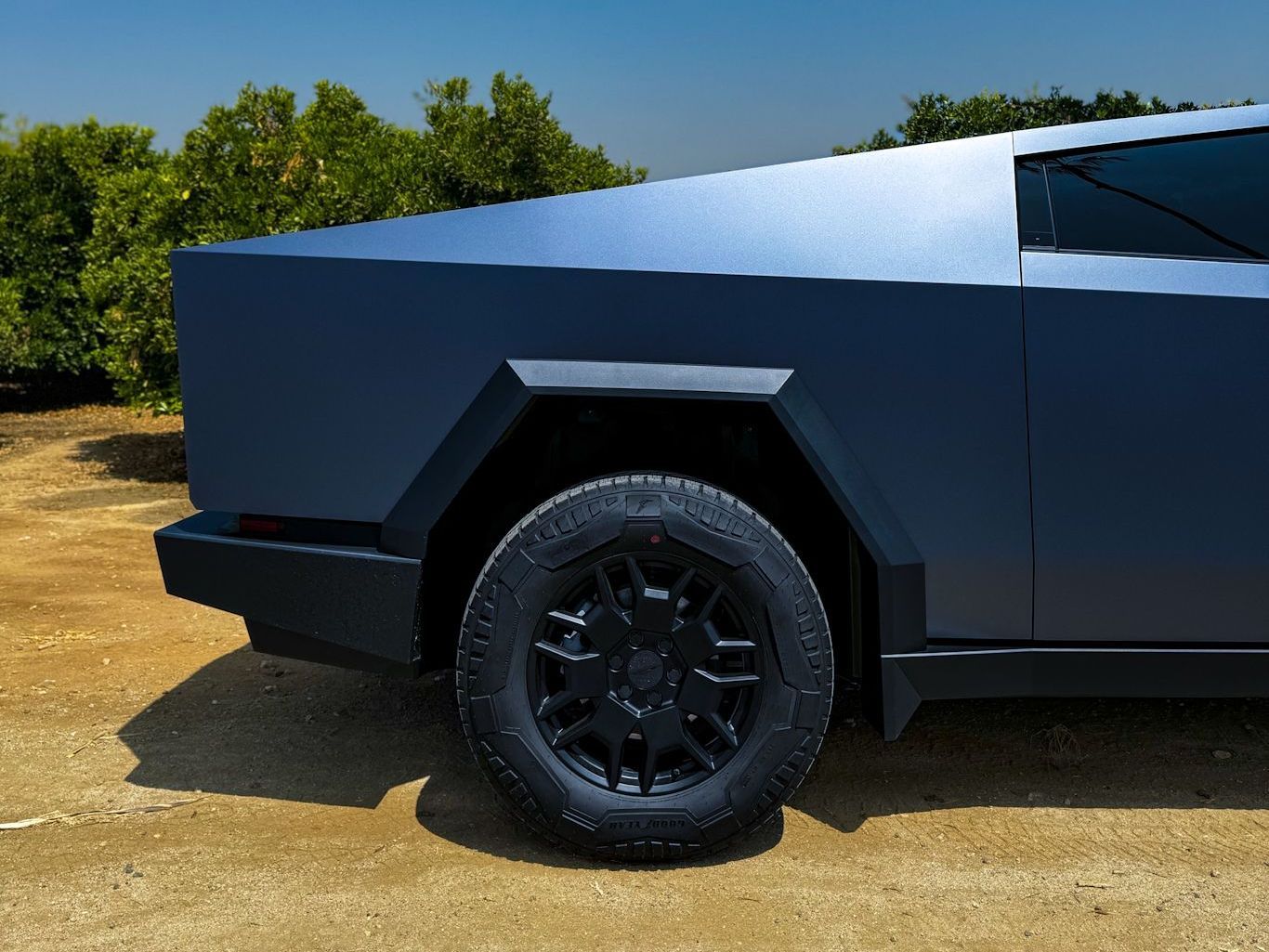 A blue truck is parked in a dirt field with trees in the background.