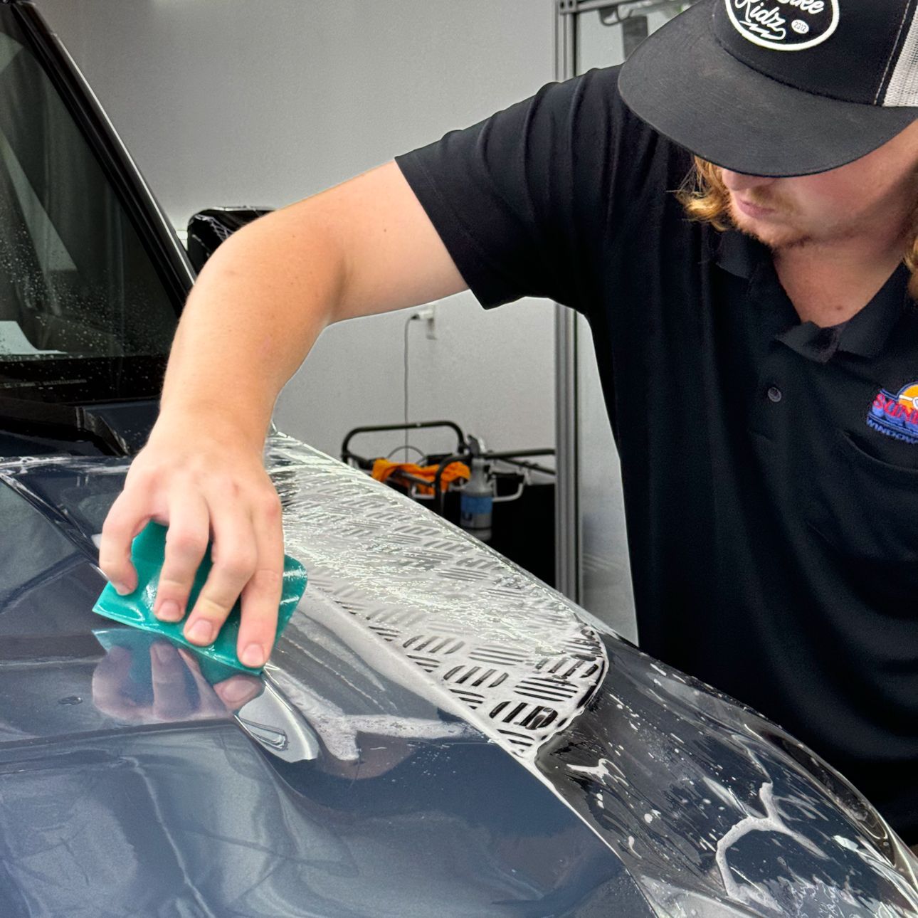 A man in a hat is applying a protective film to a car 's windshield.