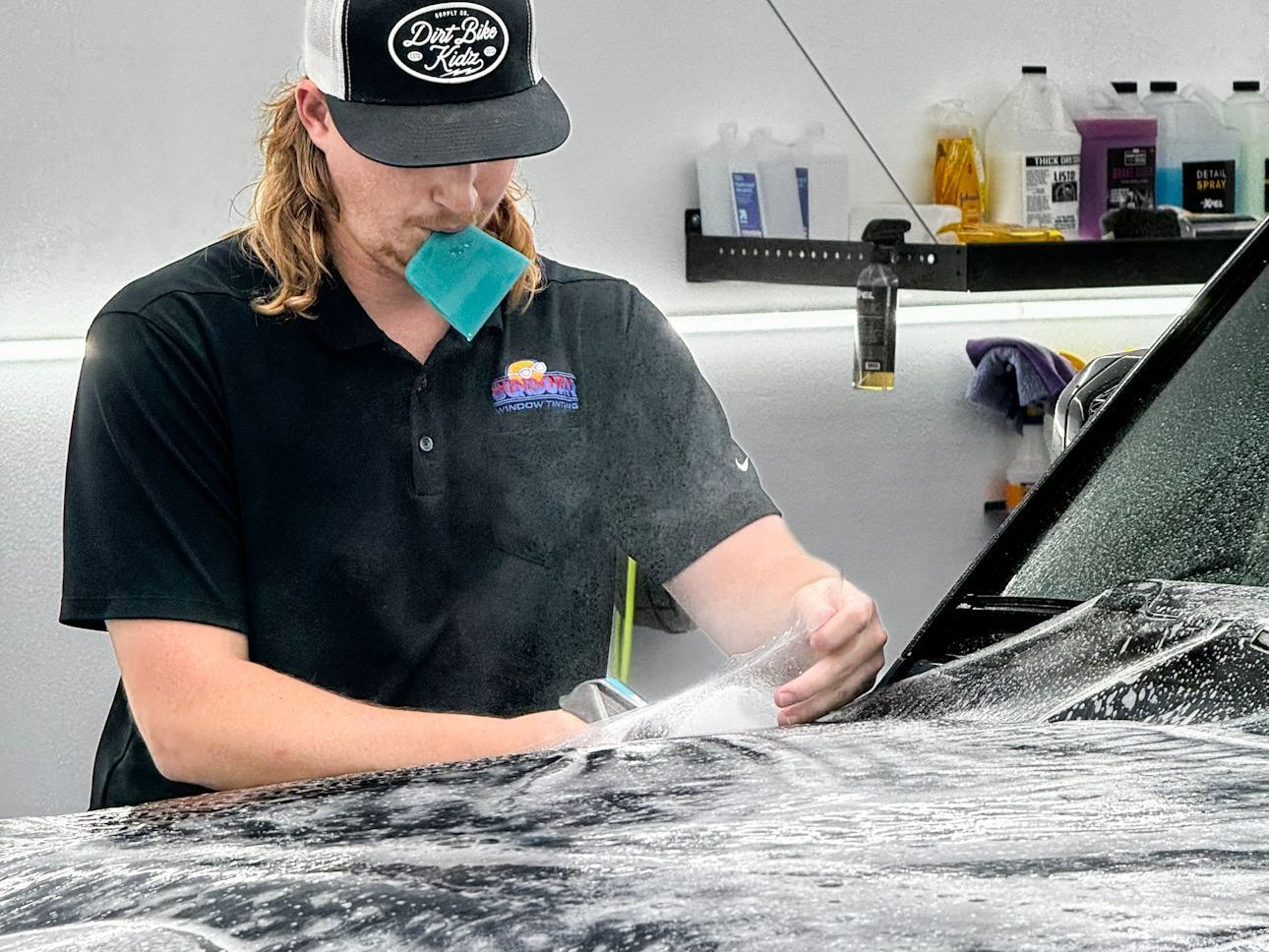 A man in a hat is applying a protective film to a car 's windshield.