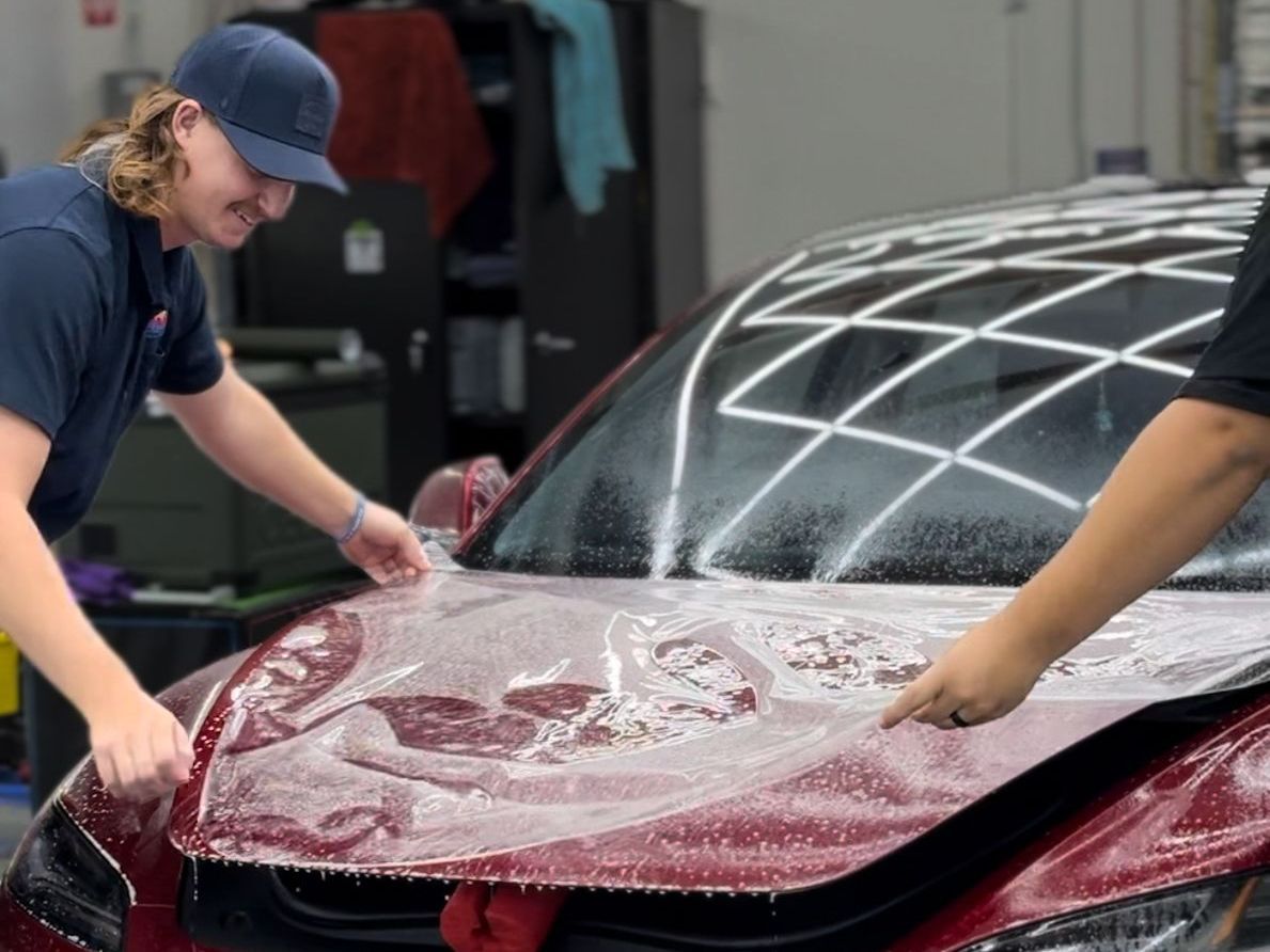 A man is covering the hood of a red car with plastic wrap