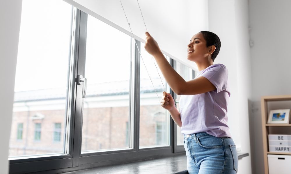 A woman is hanging blinds on a window in a living room.