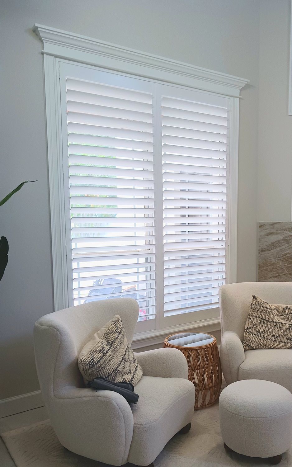 Bright living room with white shutters, beige armchair, matching ottoman, and patterned pillows