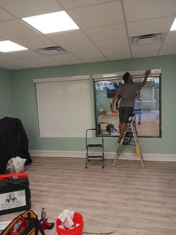 Person on a ladder painting a window in a mostly empty room with mint green walls.