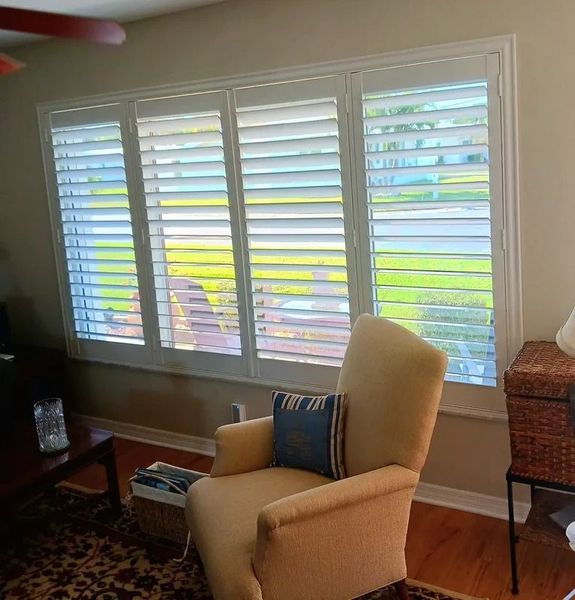 Living room with beige armchair, white shutters, and sunlight through windows