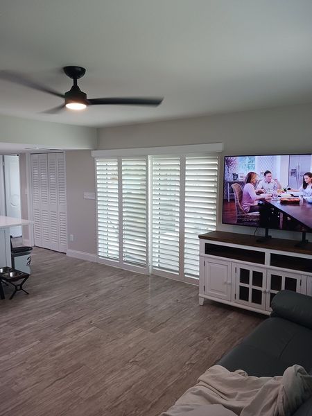 Bright living room with ceiling fan, shutters, TV, and sectional sofa on wood-look flooring.