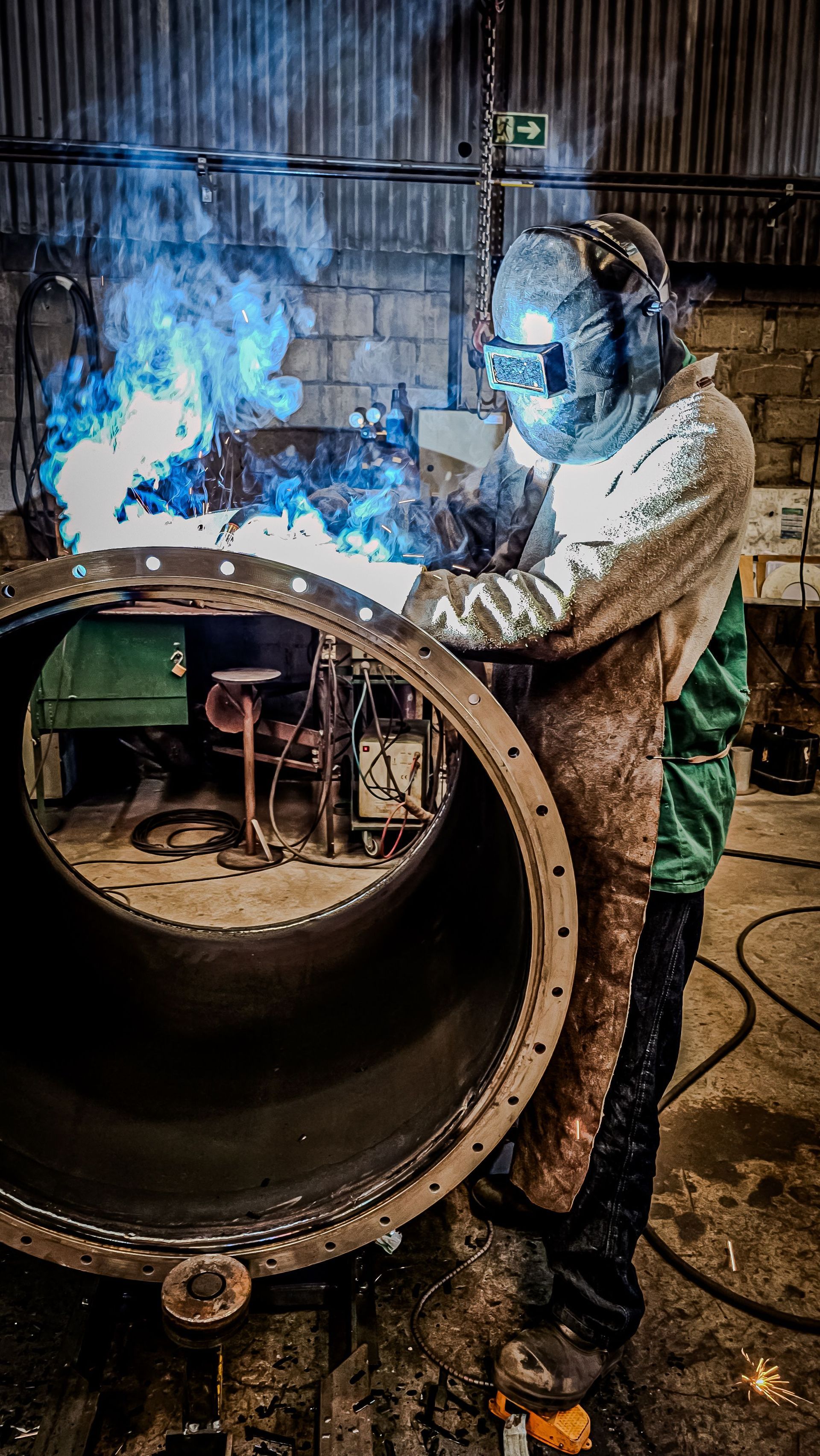 Welder in protective gear working on a large metal pipe with bright blue sparks in an industrial workshop