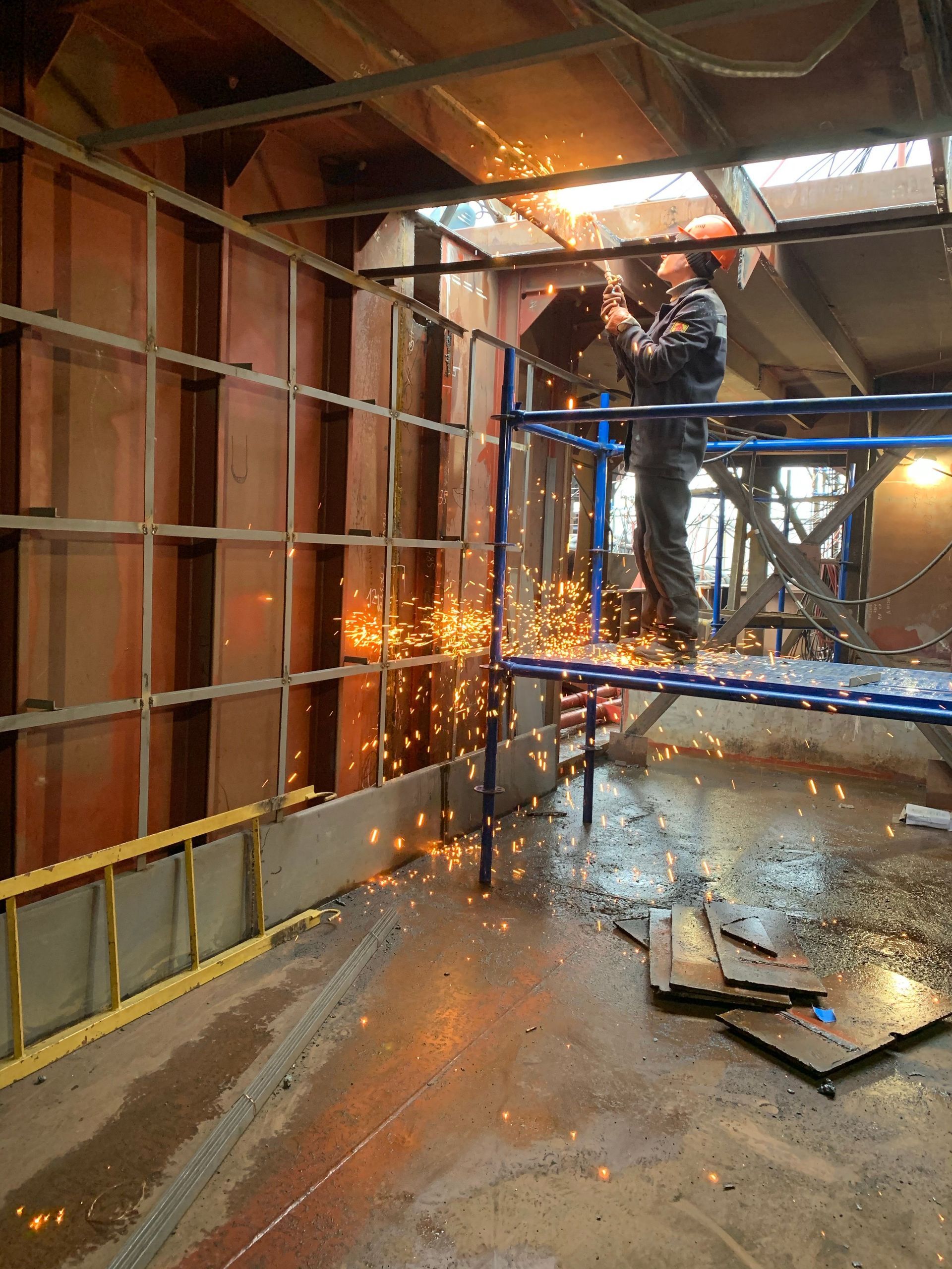 Worker cutting metal inside a narrow scaffolded corridor, with bright sparks flying and wet floor debris.
