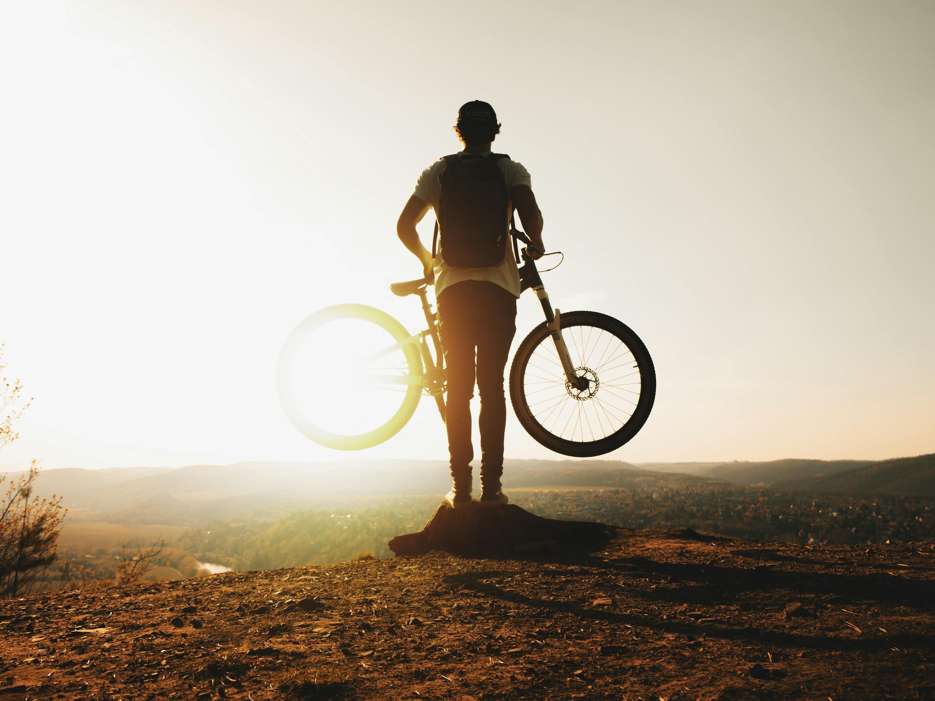 Person holding a mountain bike, silhouetted against a bright sunset on a hilltop.
