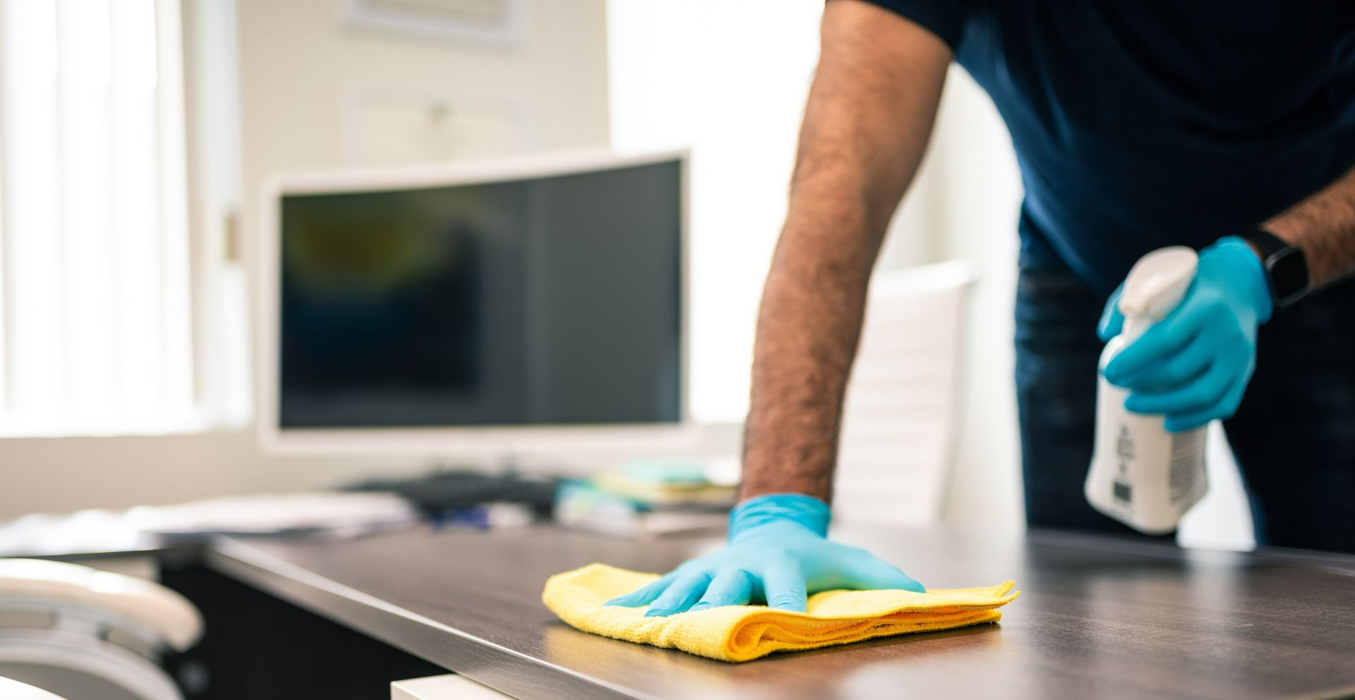 A man wearing blue gloves is cleaning a desk with a cloth and spray bottle.