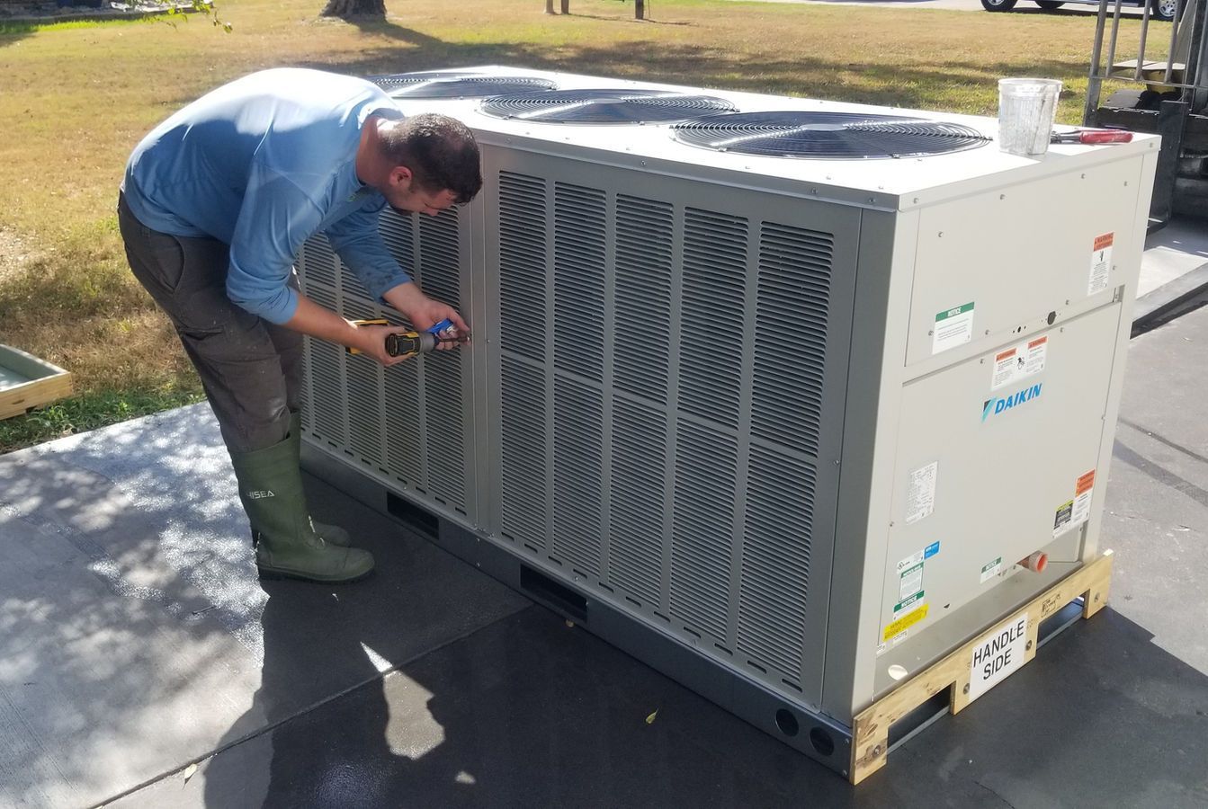 Man in blue shirt works on an air conditioning unit on a pallet outdoors.