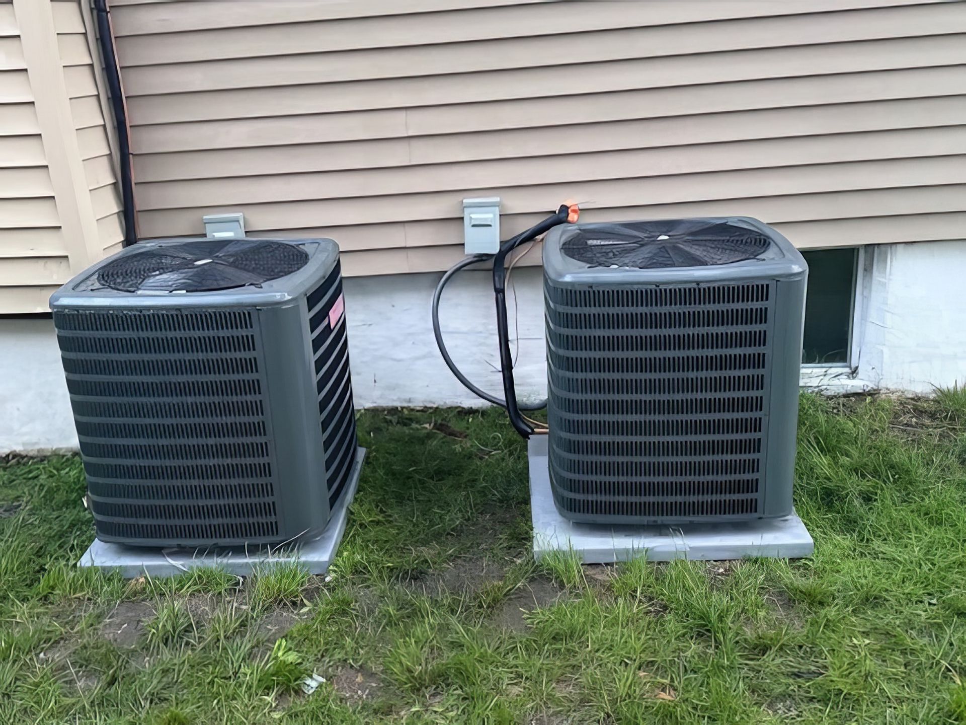 Two gray air conditioning units sitting on concrete pads next to a beige building.