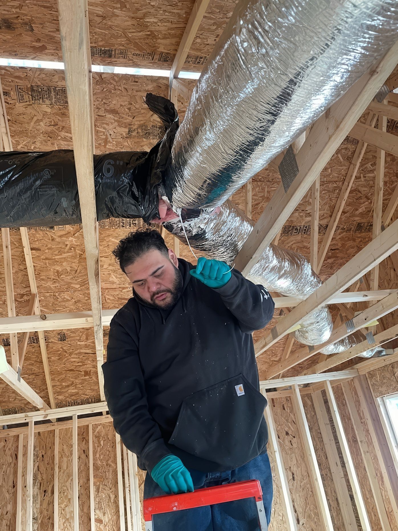 Man in black hoodie, blue gloves working on HVAC ductwork in a wood-framed house under construction.