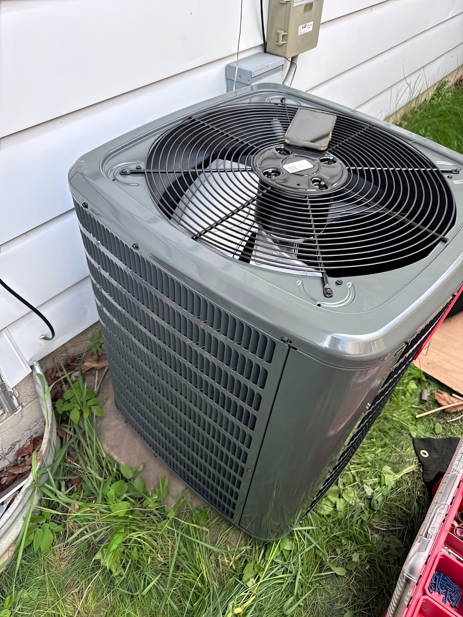 Gray air conditioning unit outside a white house on a concrete pad with a dark fan and black vents.