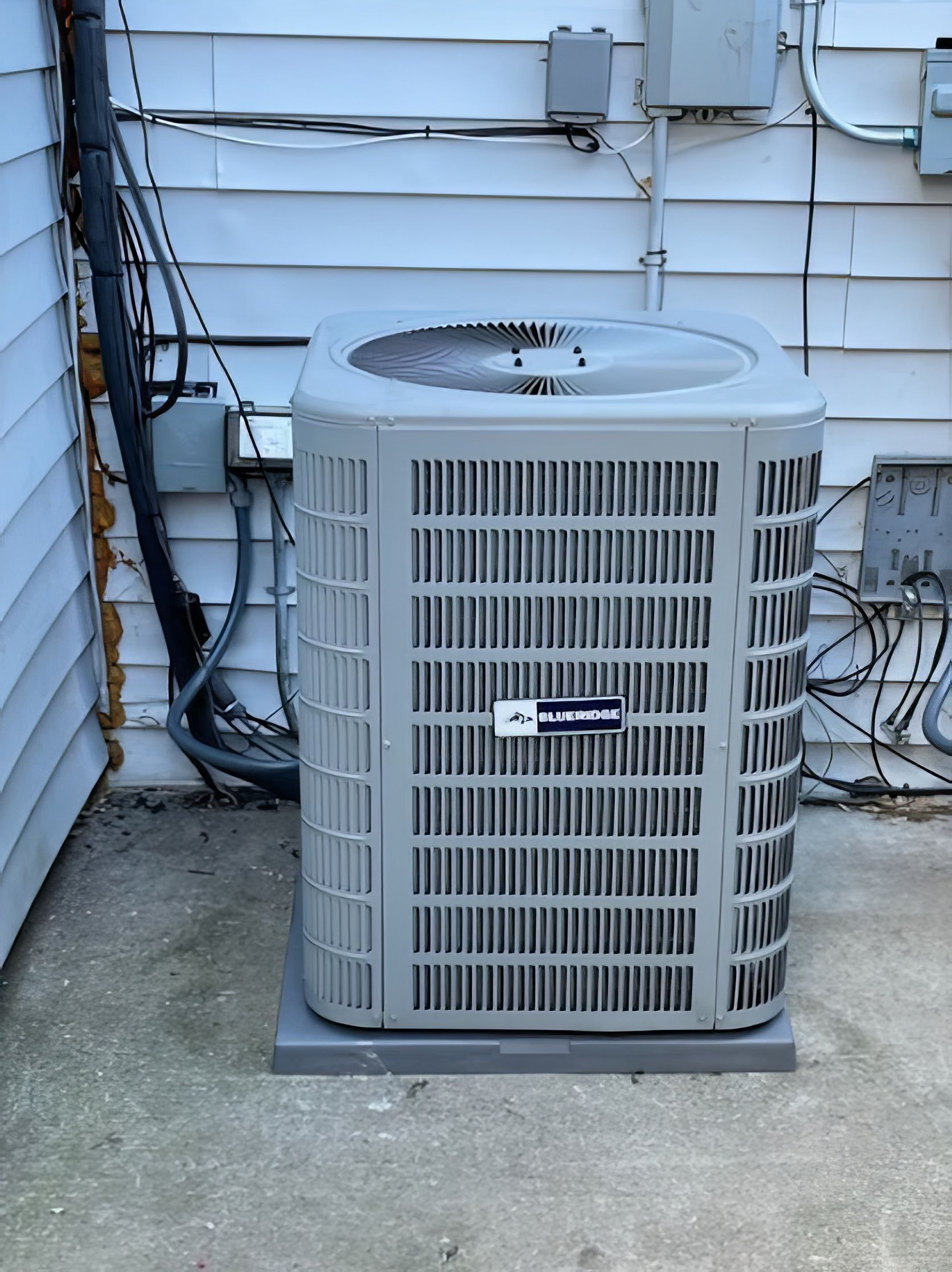 Air conditioning unit on a concrete pad next to a light-colored building wall. Electrical boxes and conduit visible.