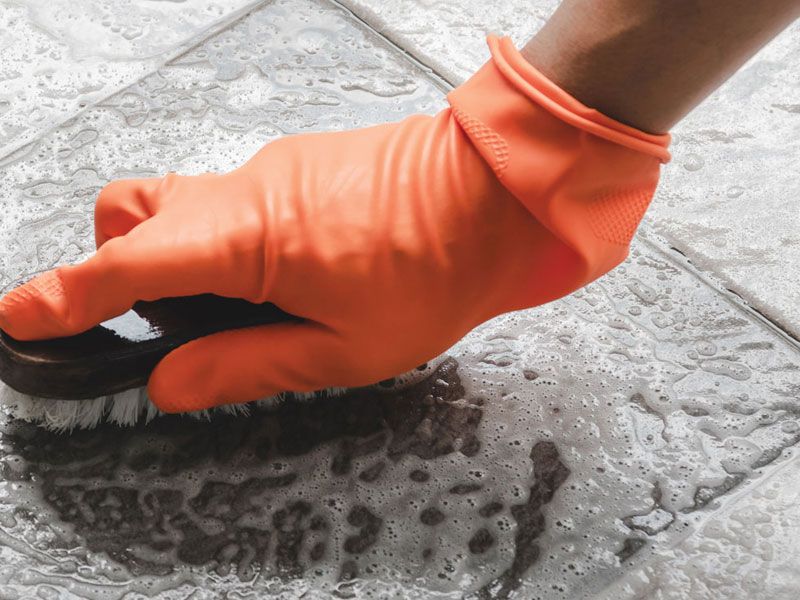 A person wearing orange rubber gloves is cleaning a tile floor with a brush