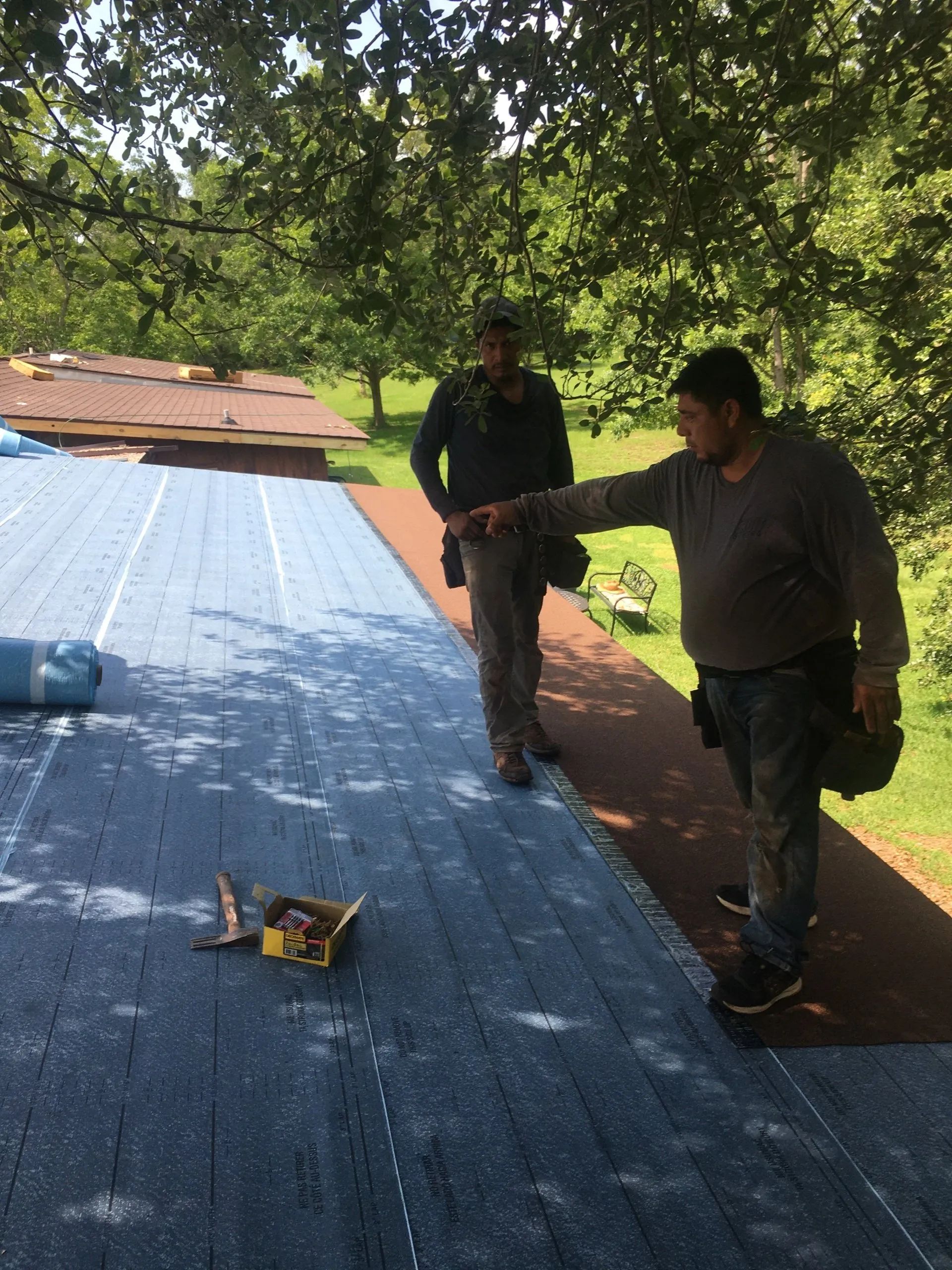 Two workers inspect a blue roof under trees, with tools and materials nearby.