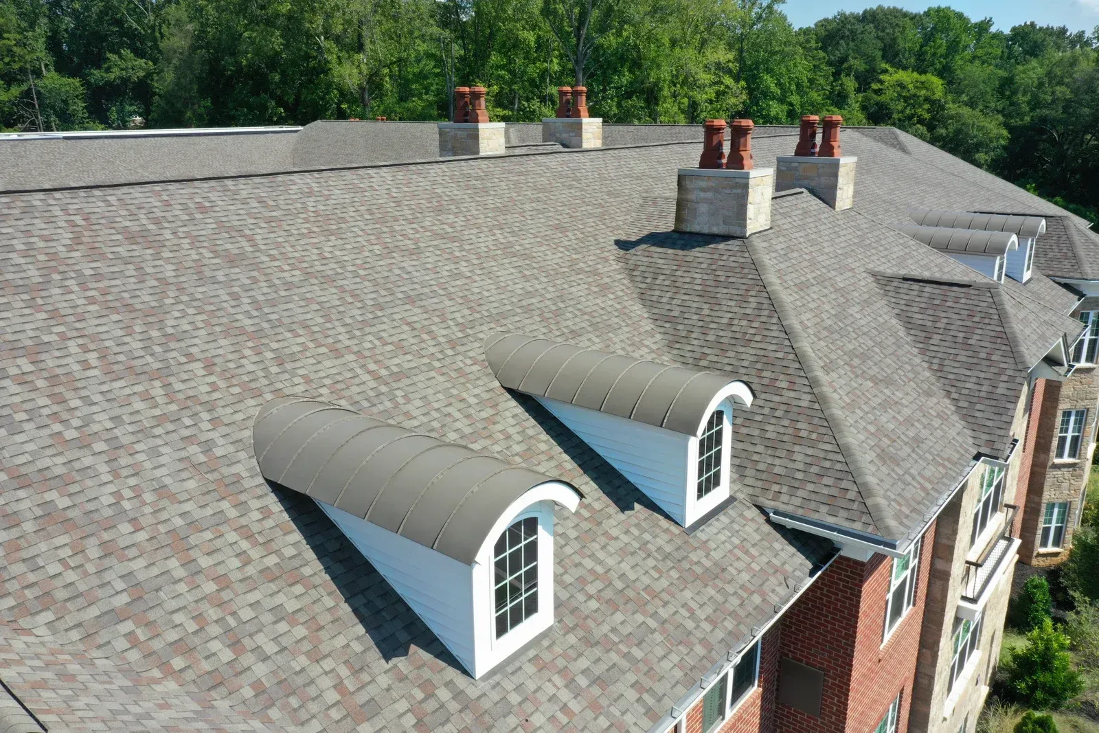 Brown shingled roof of a building with dormer windows and chimneys, trees in background.