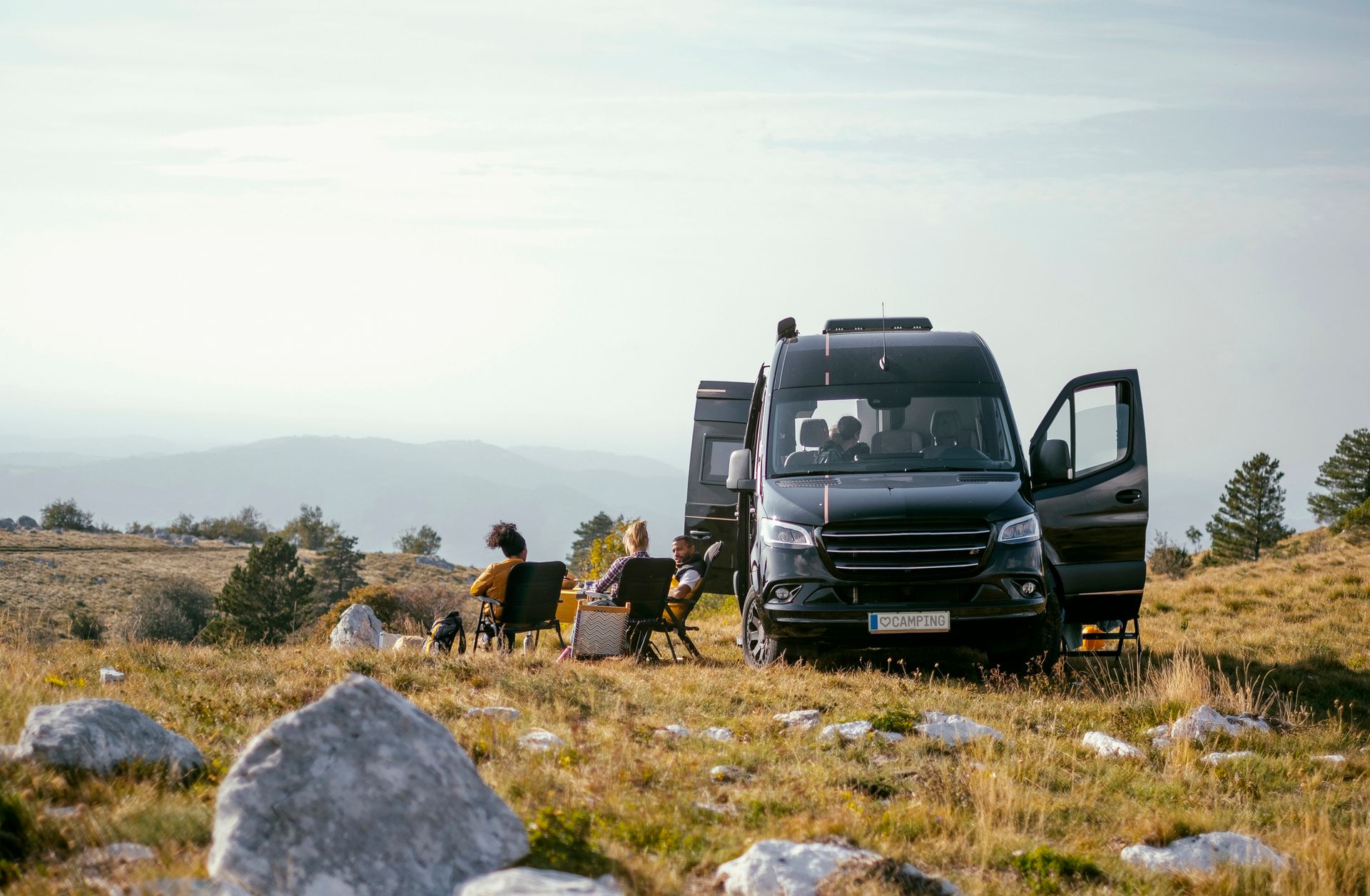 A couple of people are sitting in chairs in front of a van parked on top of a hill.