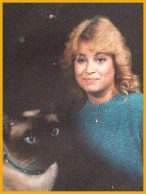 Woman with blonde hair and blue sweater smiles beside Siamese cat.