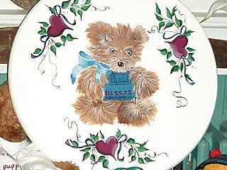 A painted teddy bear on a white plate, adorned with hearts and greenery, blue bow.