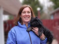 Woman in blue jacket smiling, holding small black dog outside near a red fence.