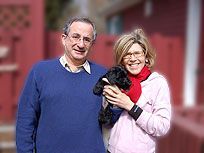 Man and woman with glasses holding a small black dog, standing in front of a red building; woman smiling.